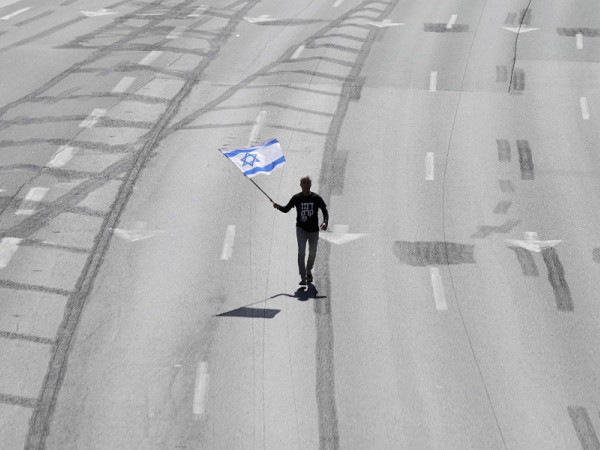 Tel Aviv (Israele). Mentre il Primo Ministro israeliano Benjamin Netanyahu parte per una visita di Stato in Italia, un manifestante sventola una bandiera israeliana lungo l’autostrada principale. Chi protesta si oppone al piano di riforma del sistema giudiziario proposto dal Governo che mira a indebolire l’autorità della Corte suprema.