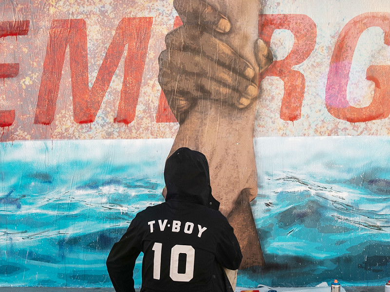 Due braccia che si afferrano tra le onde, con una scritta che spunta sullo sfondo e dice "Emerge": il murale di TvBoy realizzato per la Life Support.