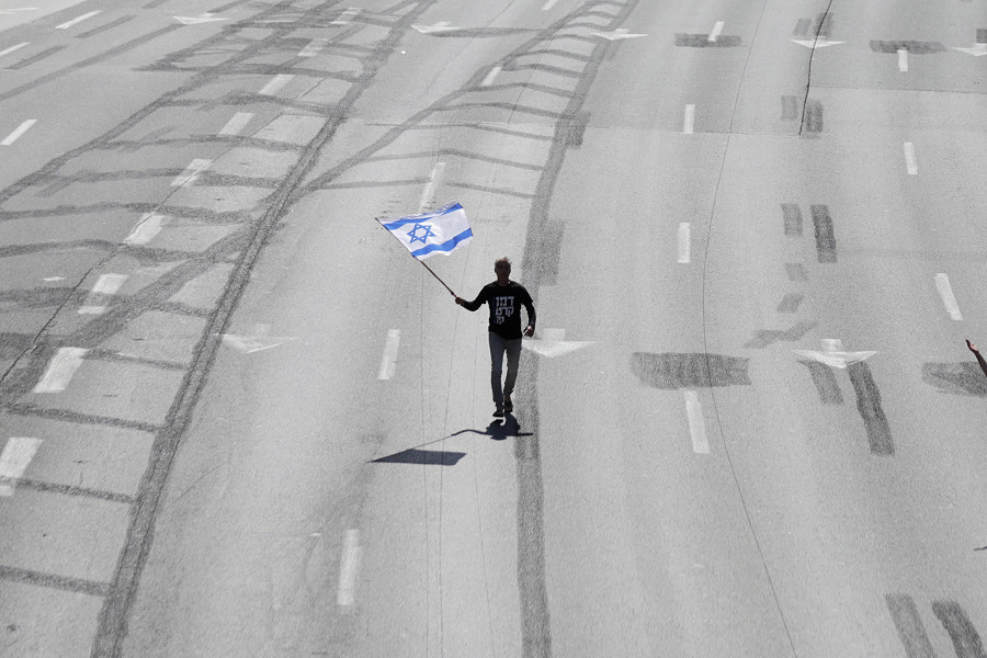 Tel Aviv (Israele). Mentre il Primo Ministro israeliano Benjamin Netanyahu parte per una visita di Stato in Italia, un manifestante sventola una bandiera israeliana lungo l’autostrada principale. Chi protesta si oppone al piano di riforma del sistema giudiziario proposto dal Governo che mira a indebolire l’autorità della Corte suprema. 
