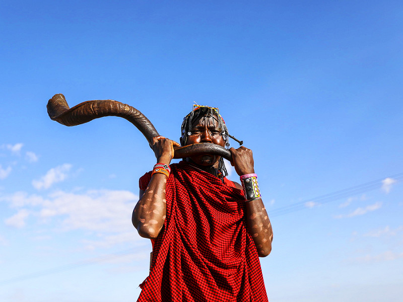 Sekenani (Kenya). Un membro della comunità Maasai in abiti tradizionali suona il corno al Festival Culturale Maasai nella Riserva Nazionale Mara. Il Festival promuove il turismo, lo scambio culturale e la pace attraverso musica, cibo, manufatti, sport tradizionali come il lancio della lancia e racconti popolari.